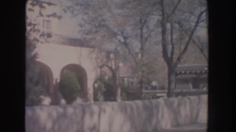 1951 New Mexico frame with pale adobe-style arches, a low stone wall, winter-bare trees, and shrubs under hazy light.