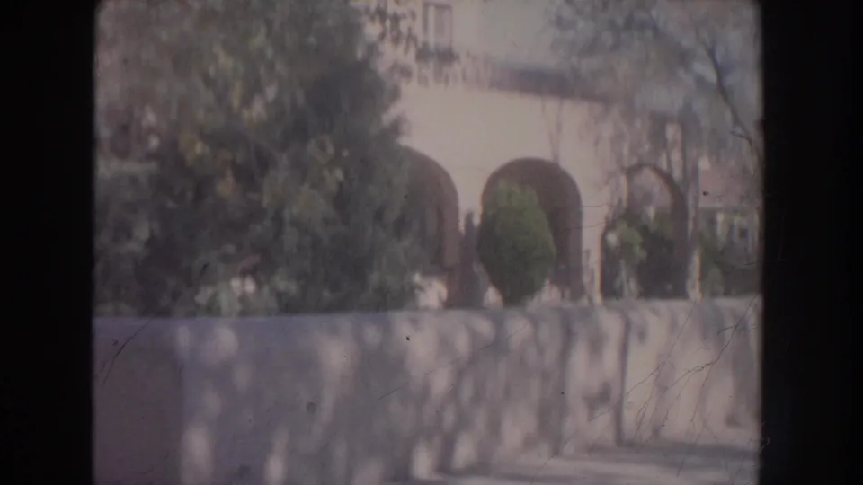 1951 New Mexico home-movie frame showing an adobe-style archway partly hidden by foliage and a stone wall.