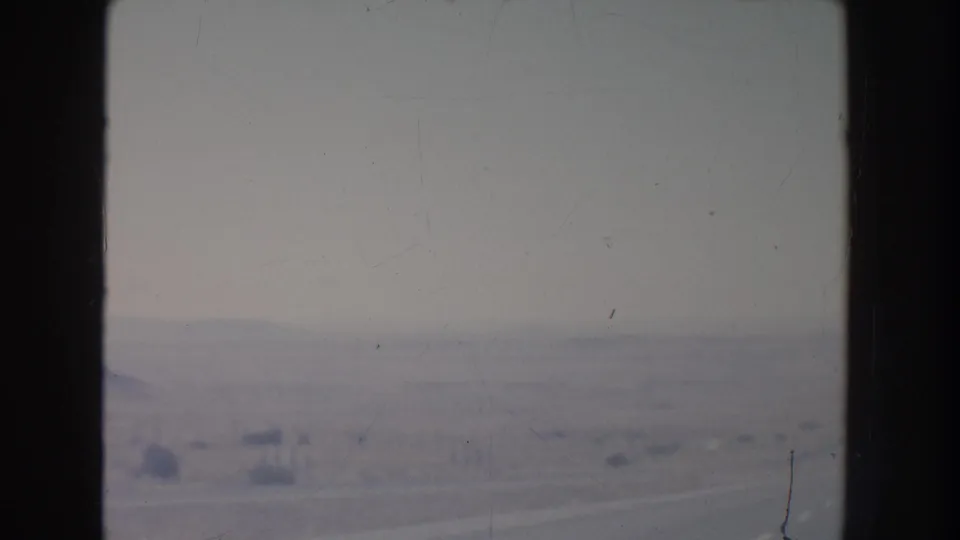 New Mexico desert in 1951 appears beyond a vehicle window, with a faint road line, low brush, and distant hills.