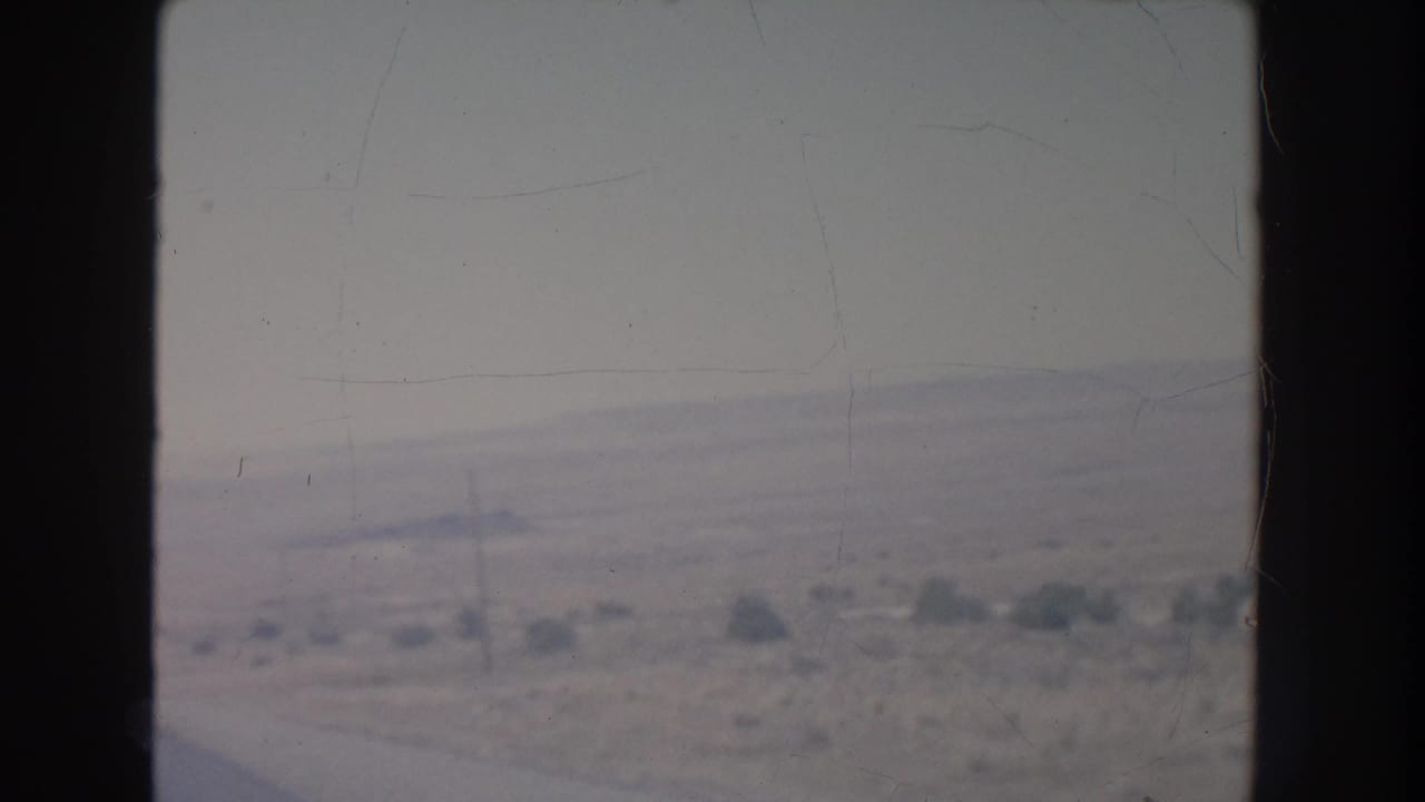Hazy desert roadside seen from a moving vehicle, with scrub and utility poles beside a pale highway.