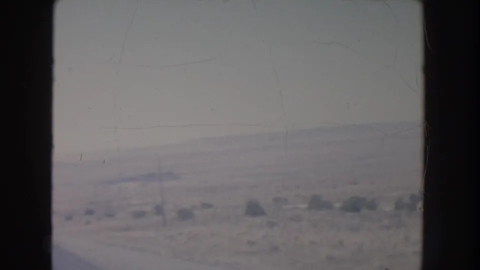 1951 New Mexico home-movie frame looking across a snowy or washed-out desert roadside from a moving vehicle, with utility poles and low scrub visible.