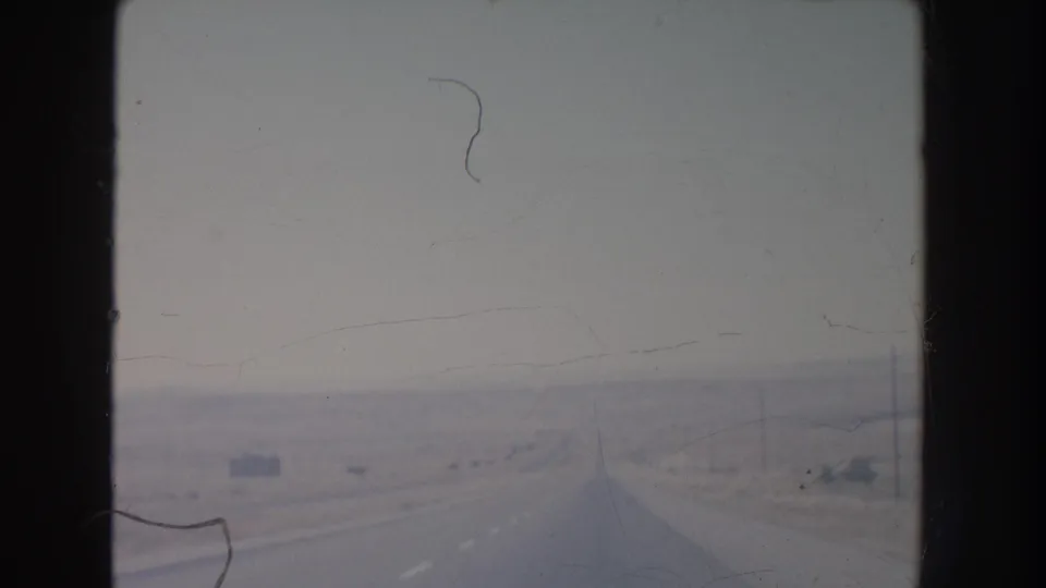 1951 New Mexico driving view down a straight, nearly empty highway through pale desert terrain under a muted sky.