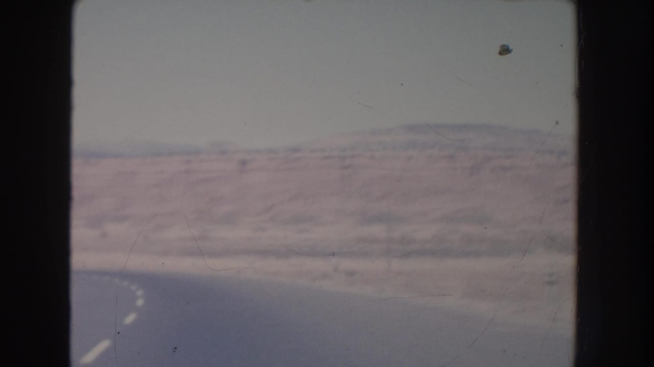 Curving desert road with mesas and distant mountains under a pale sky.