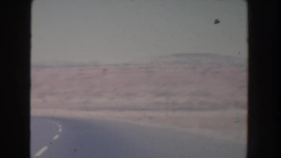 1951 New Mexico home-movie frame showing a paved road curving through desert with reddish mesas and distant mountains.