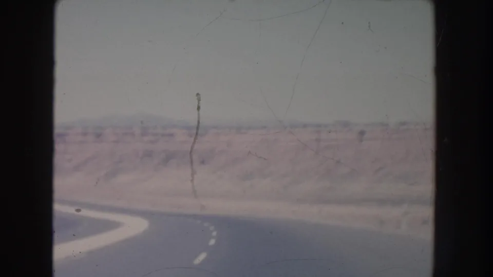 New Mexico desert road viewed from a moving vehicle, with a pale curve of pavement and mesa landforms in the distance.