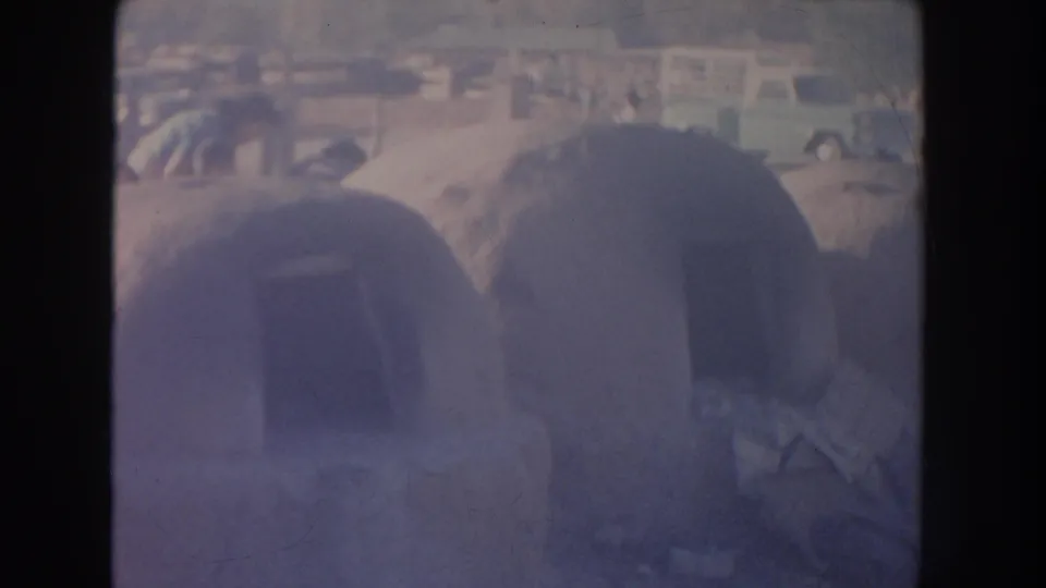 Faded 1951 New Mexico 8mm frame showing rounded adobe structures near a dusty roadside with older cars beyond.