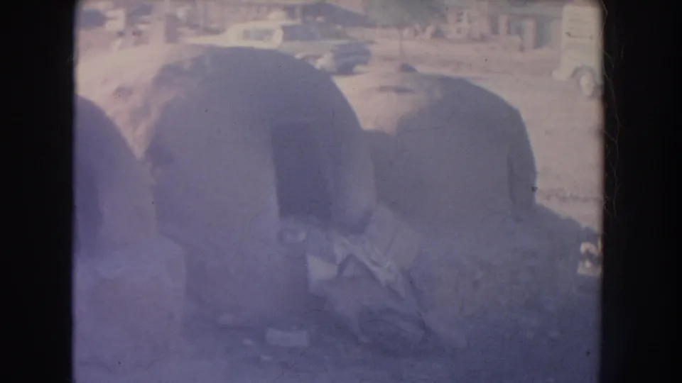 1951 New Mexico home-movie frame showing people near rounded adobe structures, with cars and a street visible in the background.