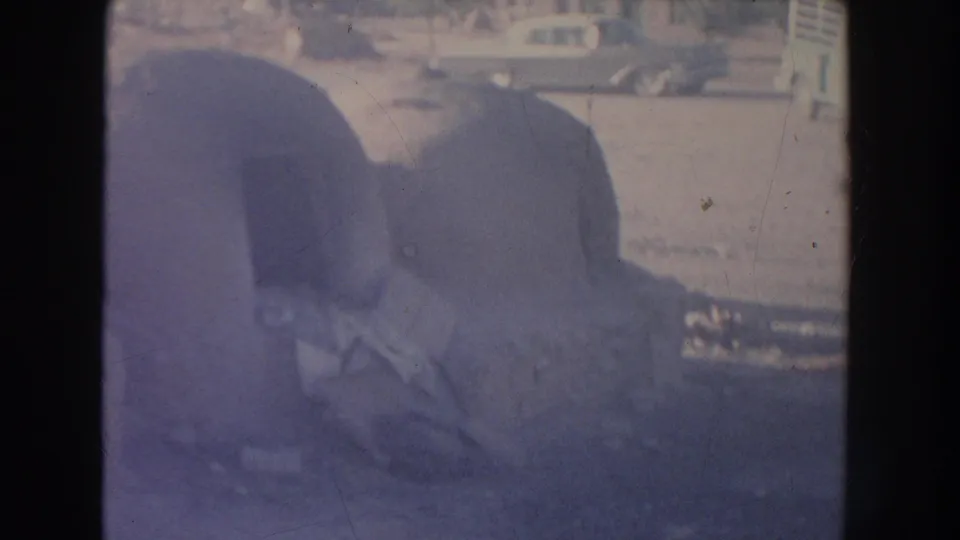 Faded 1951 New Mexico frame showing rounded adobe structures by a street, with old cars and a partially visible person nearby.