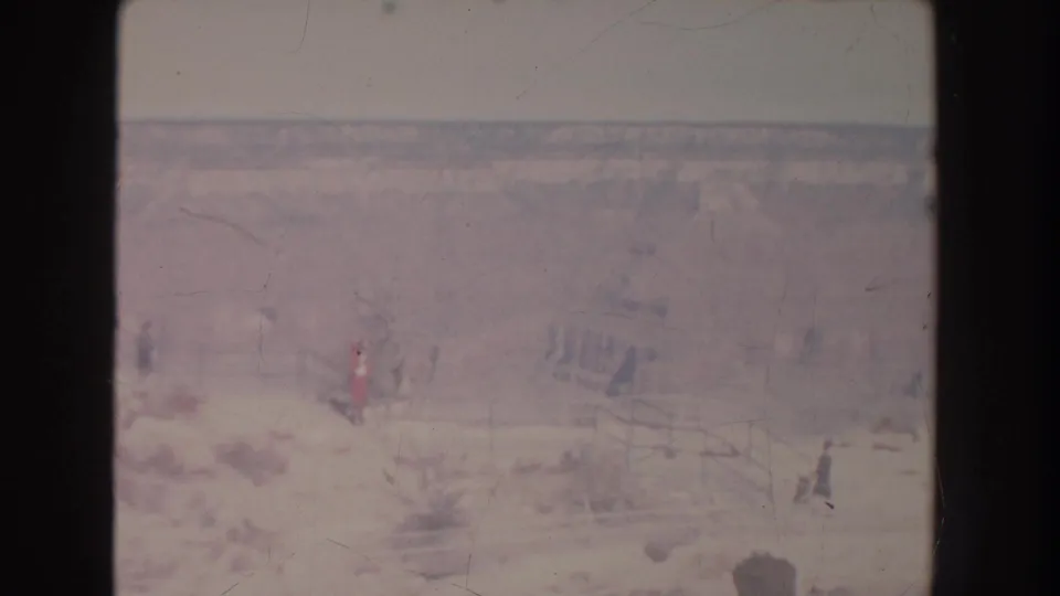 1951 New Mexico home-movie frame showing small figures on a rocky canyon path with a wooden safety railing and hazy canyon walls beyond.