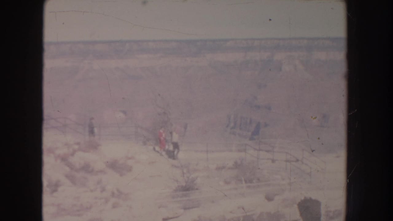 1951 New Mexico frame with several distant visitors near a wooden-railed path, set against a broad, faded canyon landscape.