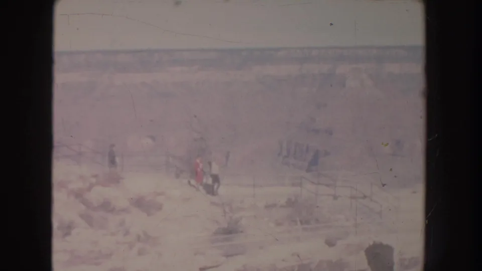1951 New Mexico frame with several distant visitors near a wooden-railed path, set against a broad, faded canyon landscape.