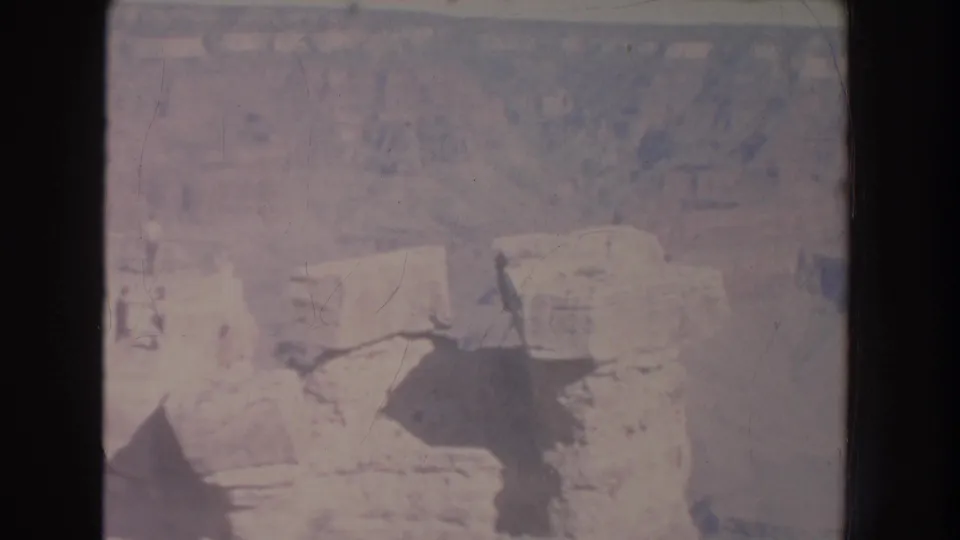 Soft 8mm frame of a rocky New Mexico canyon area with railing, scrubby vegetation, and shadowed foreground shapes from the overlook.