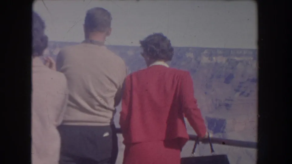 1951 New Mexico home-movie frame with an older man in a light sweater and a woman in a red suit standing at a canyon overlook rail.