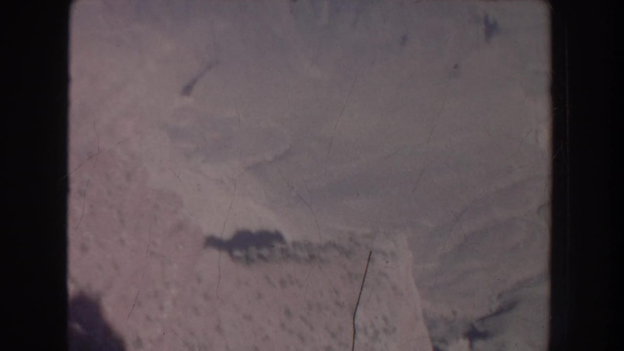 Blurred aerial landscape with dark ridge shapes, pale ground, and vertical film marks.