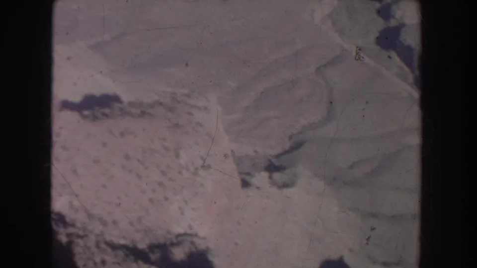 1951 New Mexico aerial frame with pale rugged terrain, dark ridge-like shapes, soft exposure, and scratches from aged 8mm film.