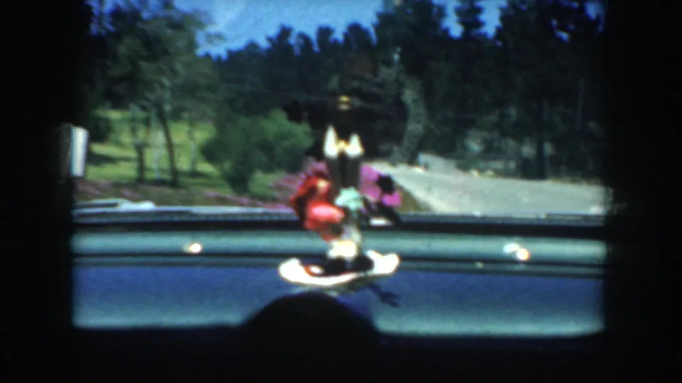 1952 California road footage seen from inside a moving car, with dashboard figurine, windshield, trees, and landscaped roadside visible.