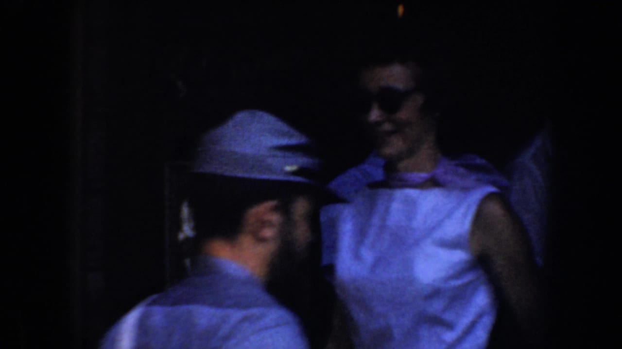 A man in a cap passes near a woman in sunglasses beside a dark entrance.