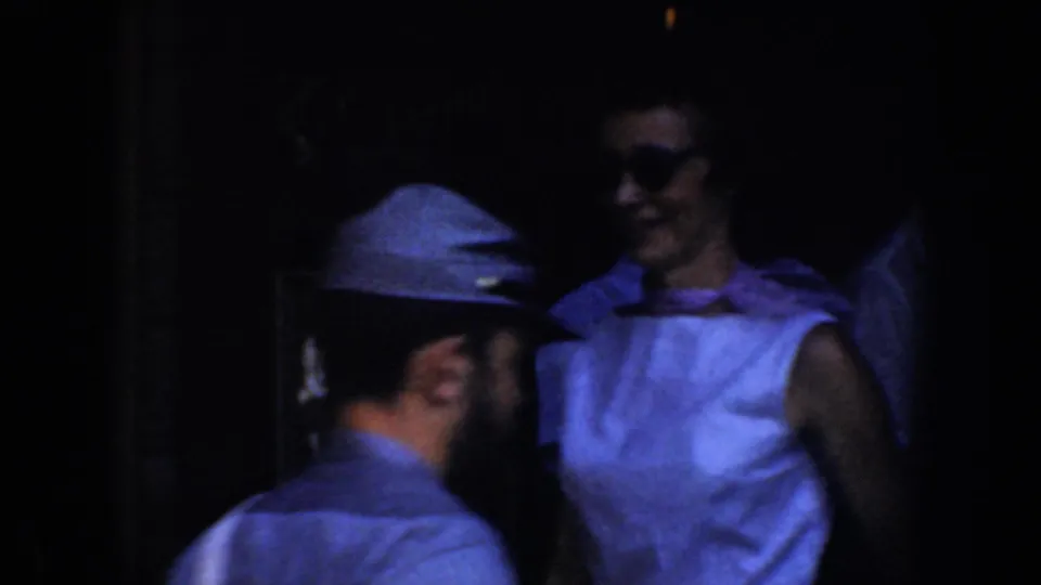 1952 California home-movie frame with a capped man and a woman in sunglasses near a dim doorway.