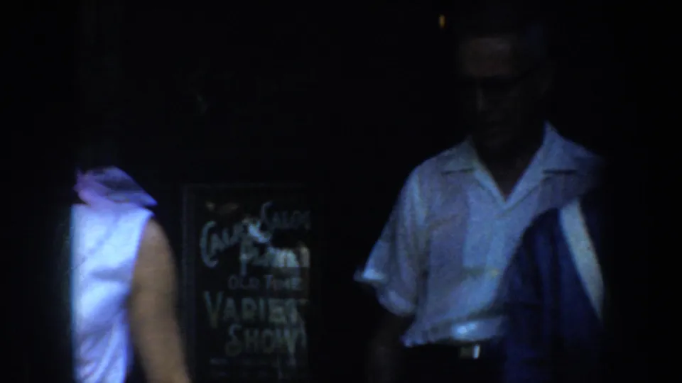 1952 California 8mm frame showing an adult near a chalkboard variety-show sign in dim light.
