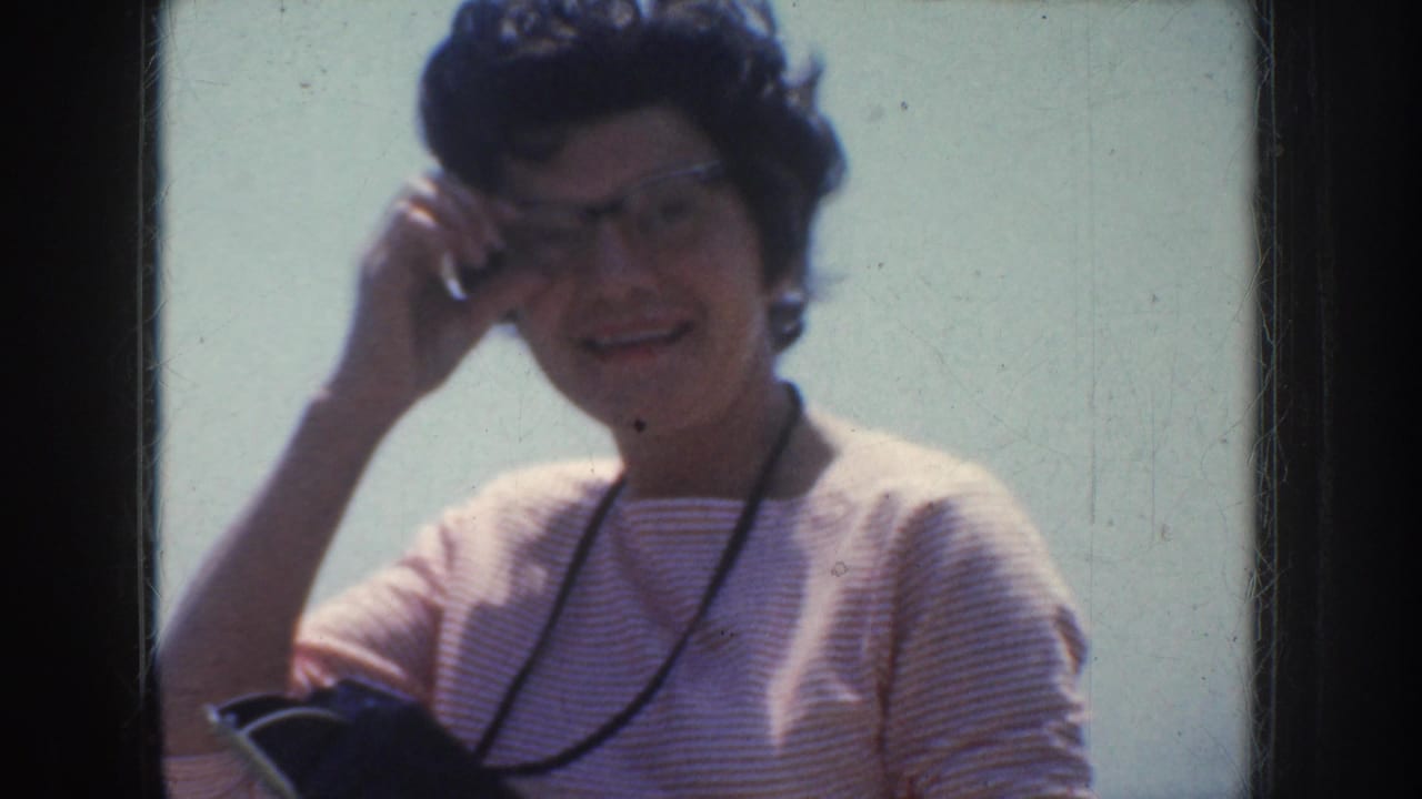 In 1952 California home-movie footage, a woman in glasses and a striped top smiles near the seaside while holding a small dark item.