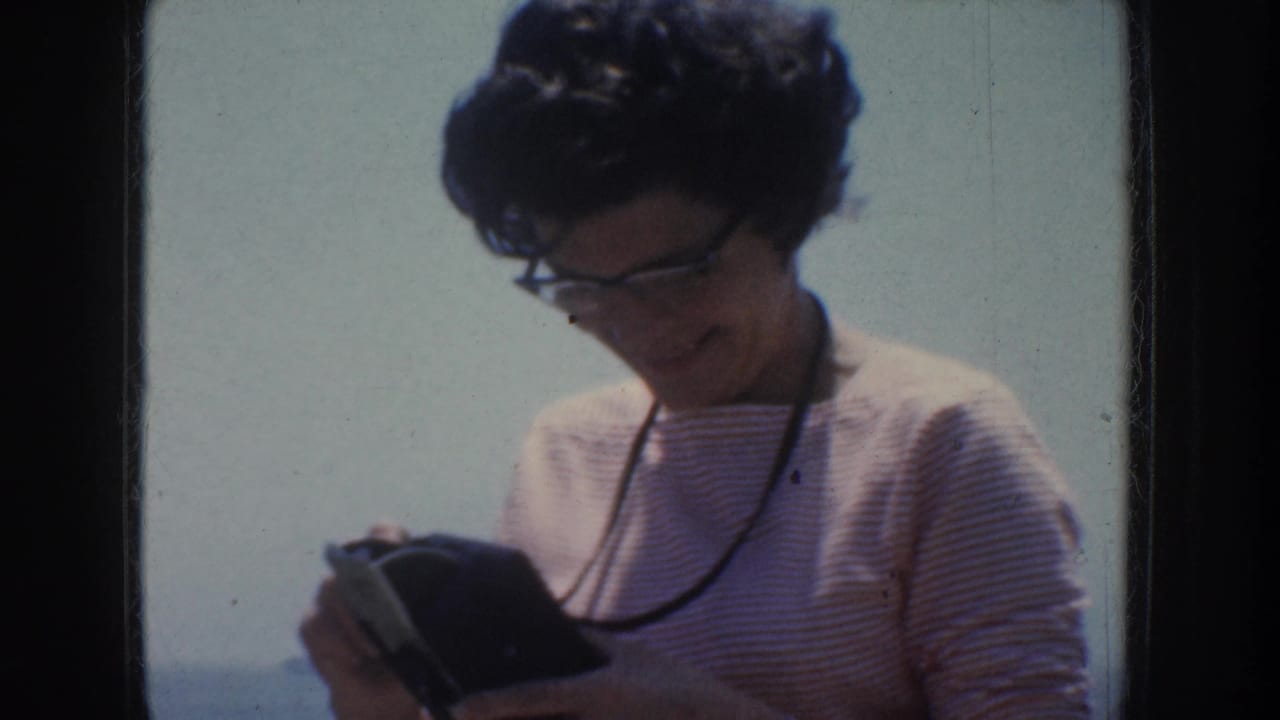Woman in glasses looking down at a small dark object against a pale sky and sea background.