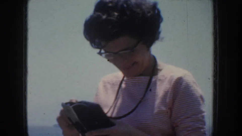 A woman in a striped top and glasses looks down at a small dark object in bright California seaside light.