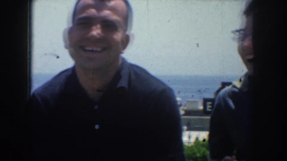 A man in a dark polo shirt smiles toward the home-movie camera with sea, sky, and partial buildings in the background.