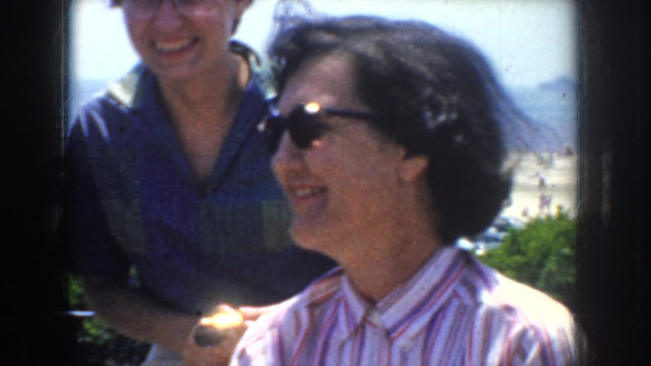 1952 California home-movie frame of two adult women smiling in bright sun, with sunglasses, casual clothing, and a hazy outdoor background.