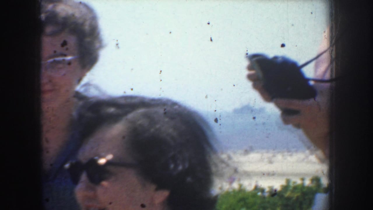Two women in sunglasses outdoors, with a hand holding a small camera at the right edge.