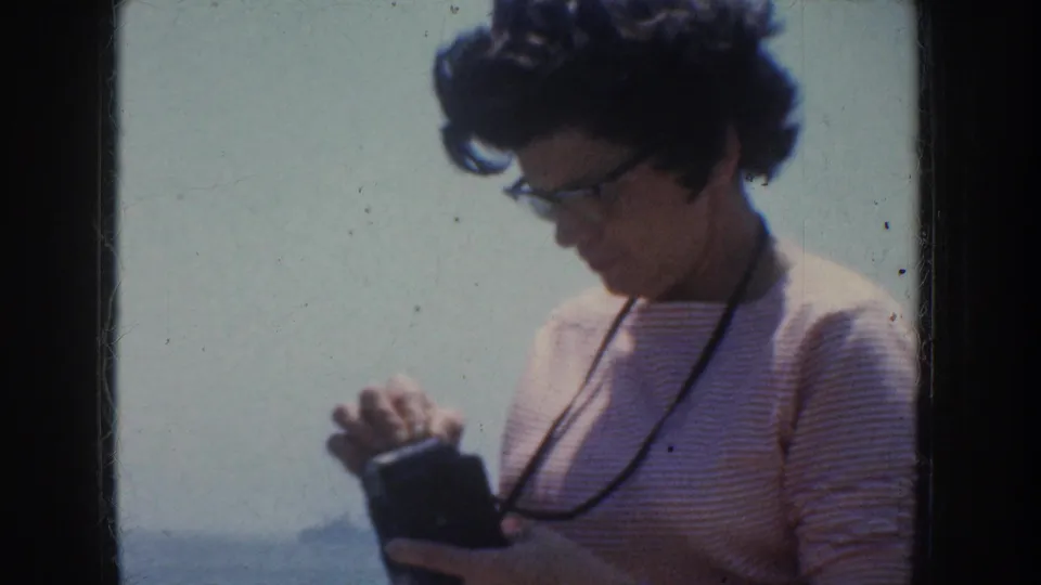 1952 California home-movie frame of an adult woman in glasses looking down at a small camera against a pale outdoor backdrop.