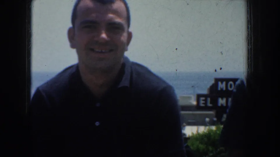 1952 California frame of an adult man smiling in a dark shirt, with distant water or haze and a partial sign behind him.