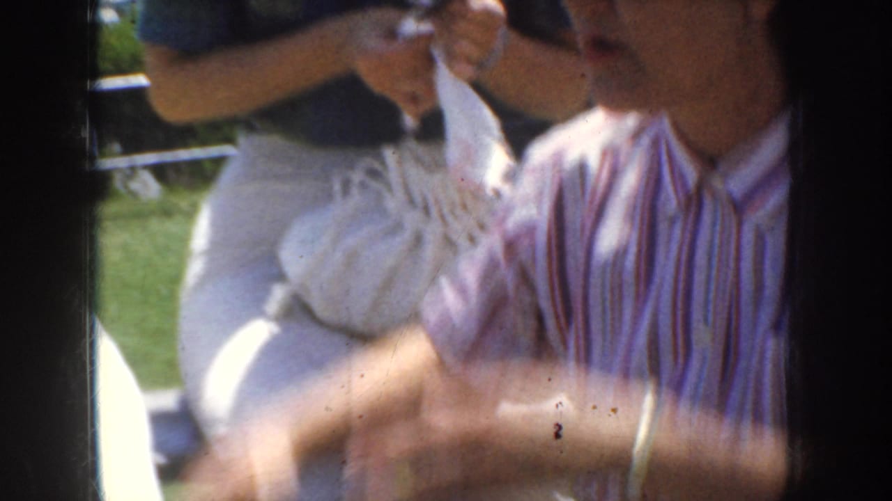 1952 California 8mm frame of a young woman in a striped shirt seated on grass beside a white drawstring bag and nearby companions.