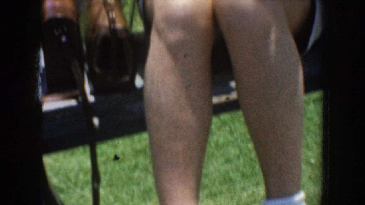 Close view of a person’s legs beside folding chairs and handbags on grass.