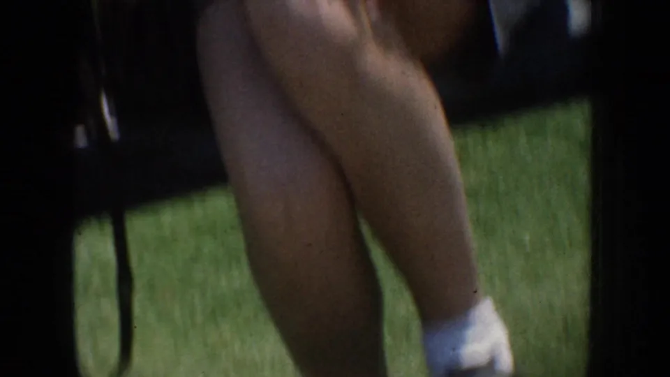 Handheld 8mm frame focused on a seated person’s legs and white socks on a sunny grass lawn during an outdoor gathering.