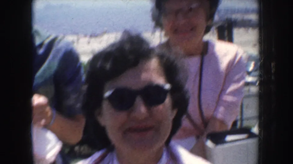 1952 California 8mm frame of a woman in dark sunglasses facing the camera, with another woman in a pink top behind her.