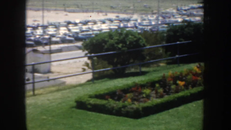 1952 California 8mm frame of a manicured garden bed, lawn, railing, and parking area seen from an overlook.