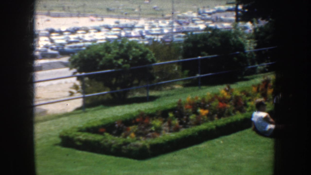 1952 California home-movie frame showing a green park lawn, flower bed, railing, distant parking area, and a seated adult at right.