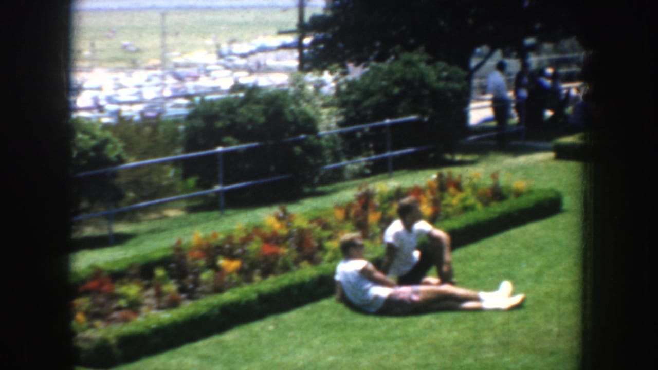 Two adults relax on a green lawn beside a flower bed, with railing and parked cars beyond.
