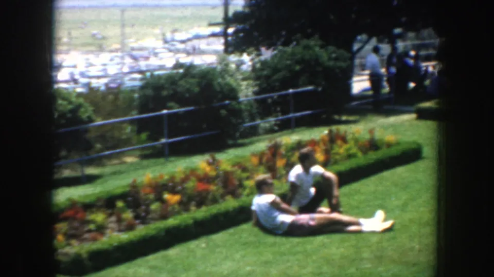 1952 California park scene with two adults resting on grass near a rectangular flower bed and a railing above a parking area.