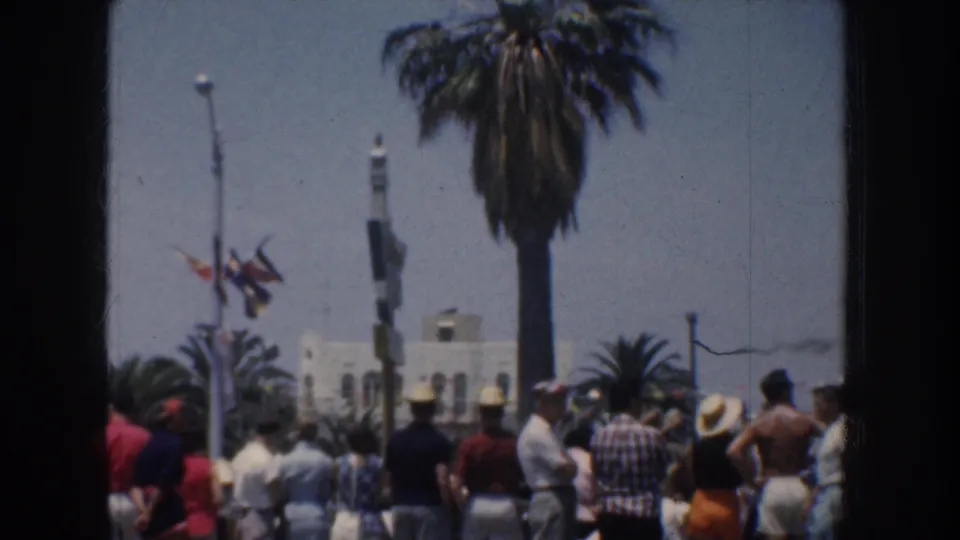 1952 California beach-resort crowd in summer clothing gathered near palms, flags, and a white building while facing the sea.