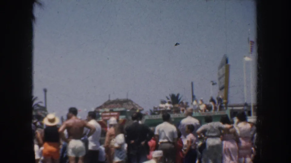 1952 California beach-resort crowd faces the ocean near palm trees, signage, and low seaside structures, with figures seen from behind.
