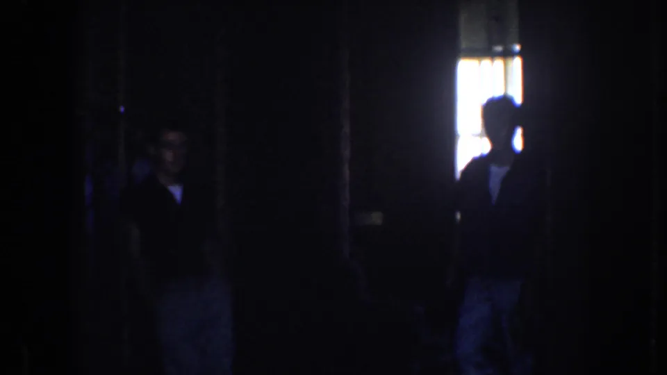 1952 California home-movie frame showing two shadowed adults near a bright window beneath chandelier details.