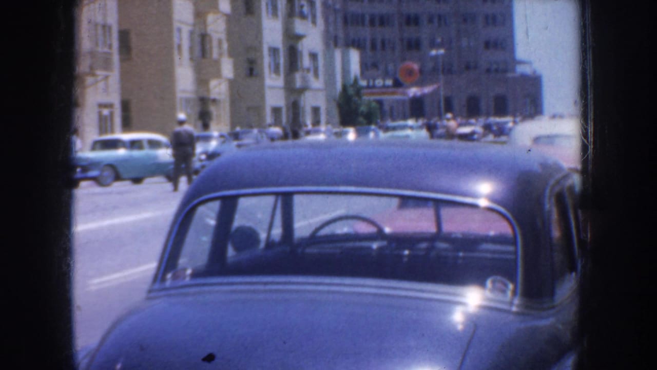 Parked dark blue sedan on a city street with buildings and cars behind it.