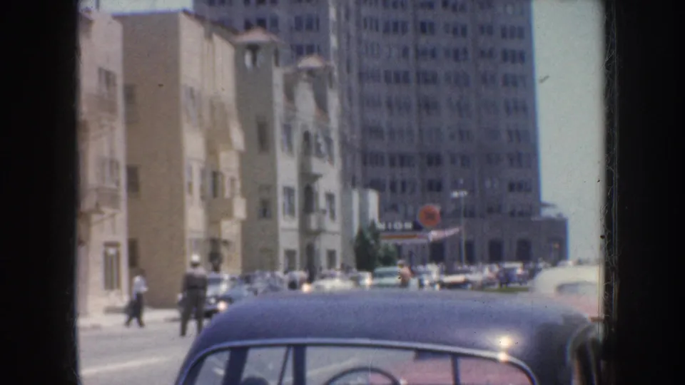 1952 California home-movie view of a city street, with a parked dark sedan in front and pedestrians crossing near tall buildings.