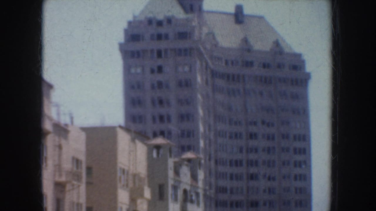 1952 California home-movie frame showing a tall downtown building with many windows and nearby low-rise structures.