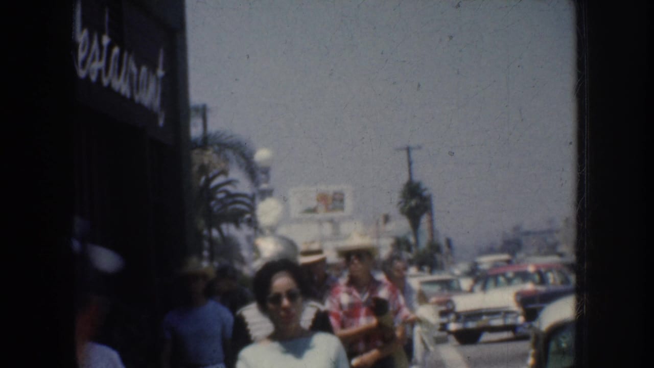 1952 California home-movie frame showing pedestrians on a sidewalk with palm trees, storefront signage, and mid-century cars in the street.