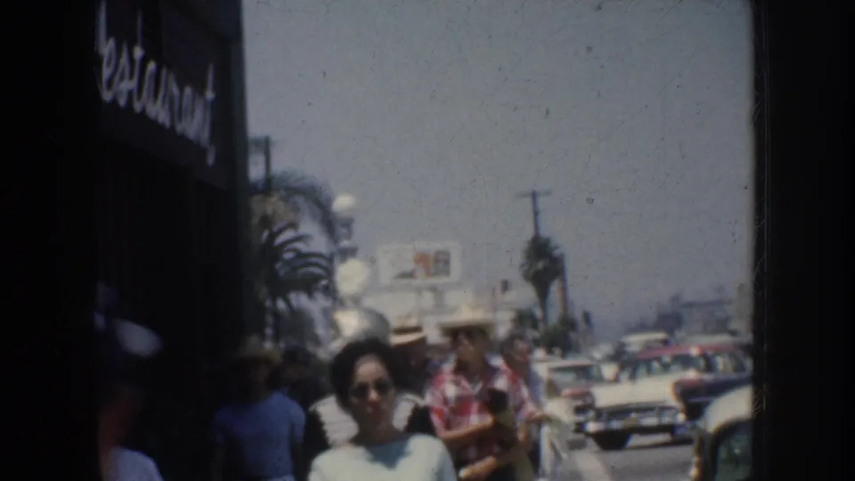 1952 California home-movie frame showing pedestrians on a sidewalk with palm trees, storefront signage, and mid-century cars in the street.