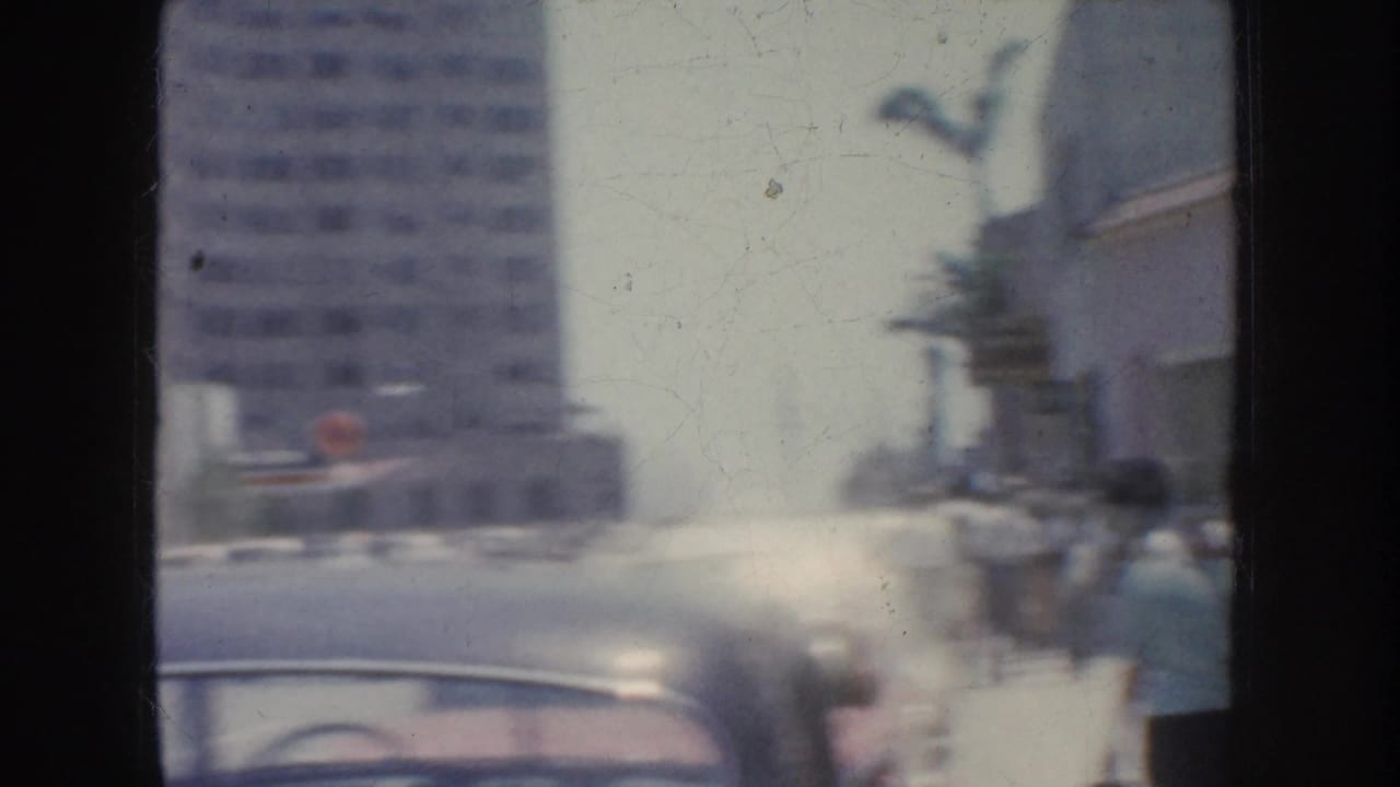 Soft 8mm street view from California in 1952, showing classic cars, pedestrians, palm trees, and multistory buildings through motion blur.