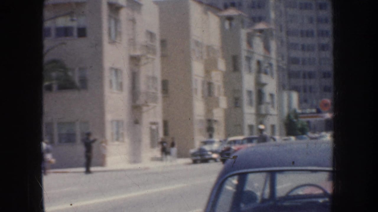 1952 California frame showing a street corner with mid-century cars, pedestrians near pale buildings, and a dark car crossing the foreground.