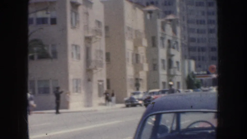 1952 California frame showing a street corner with mid-century cars, pedestrians near pale buildings, and a dark car crossing the foreground.